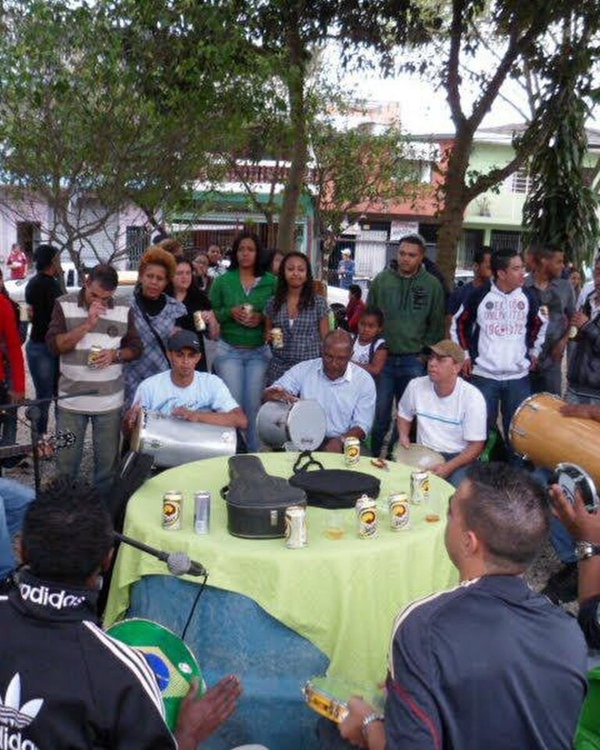 Roda de samba ao ar livre no Jardim Bussocaba, em Osasco, com músicos em volta de uma mesa, pandeiro, cavaquinho e público em pé debaixo das árvores.