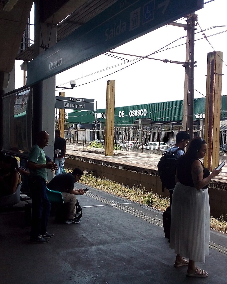 Passageiros sentados e em pé na plataforma da Estação Osasco, uma mulher olhando o celular, com o letreiro da estação e os trilhos ao fundo.