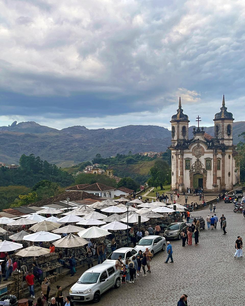 Feira de pedra-sabão do Largo do Coimbra vista do alto, com dezenas de barracas de guarda-sol branco, turistas caminhando pela rua de pedra, a Igreja de São Francisco de Assis e o Pico do Itacolomi ao fundo.
