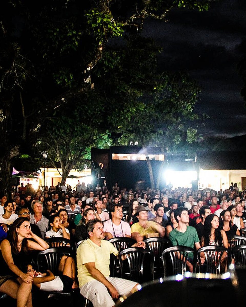 Público sentado em cadeiras ao ar livre, à noite, assistindo a uma sessão da Mostra de Cinema de Tiradentes.