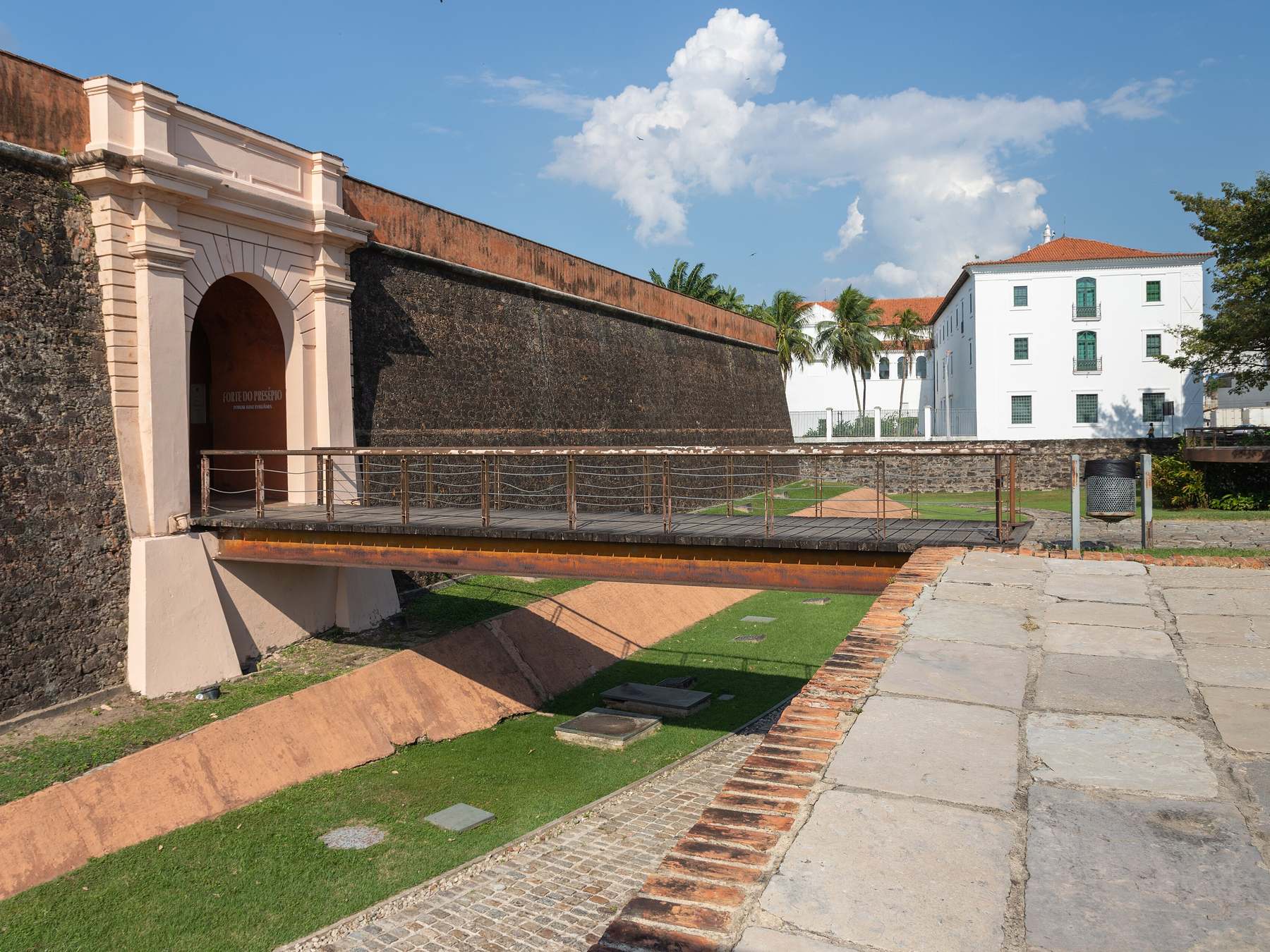 Portal de entrada e ponte de acesso do Forte do Presépio, na Cidade Velha, com muralha de pedra, um casarão colonial branco ao lado e céu azul com nuvens