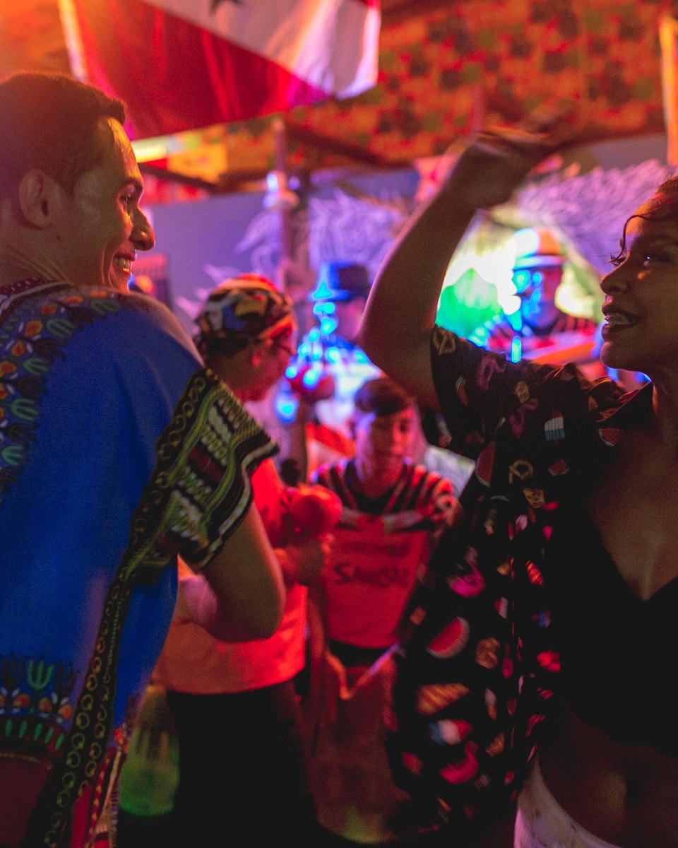 Casal dançando carimbó em roda, com os tambores e as luzes da festa ao fundo.
