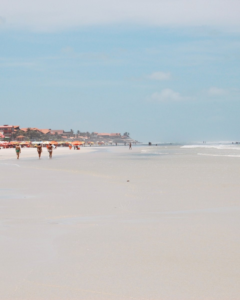 Fileira de barracas laranja e guarda-sóis na Praia do Atalaia, em Salinópolis, com carro estacionado na areia e banhistas caminhando na beira.