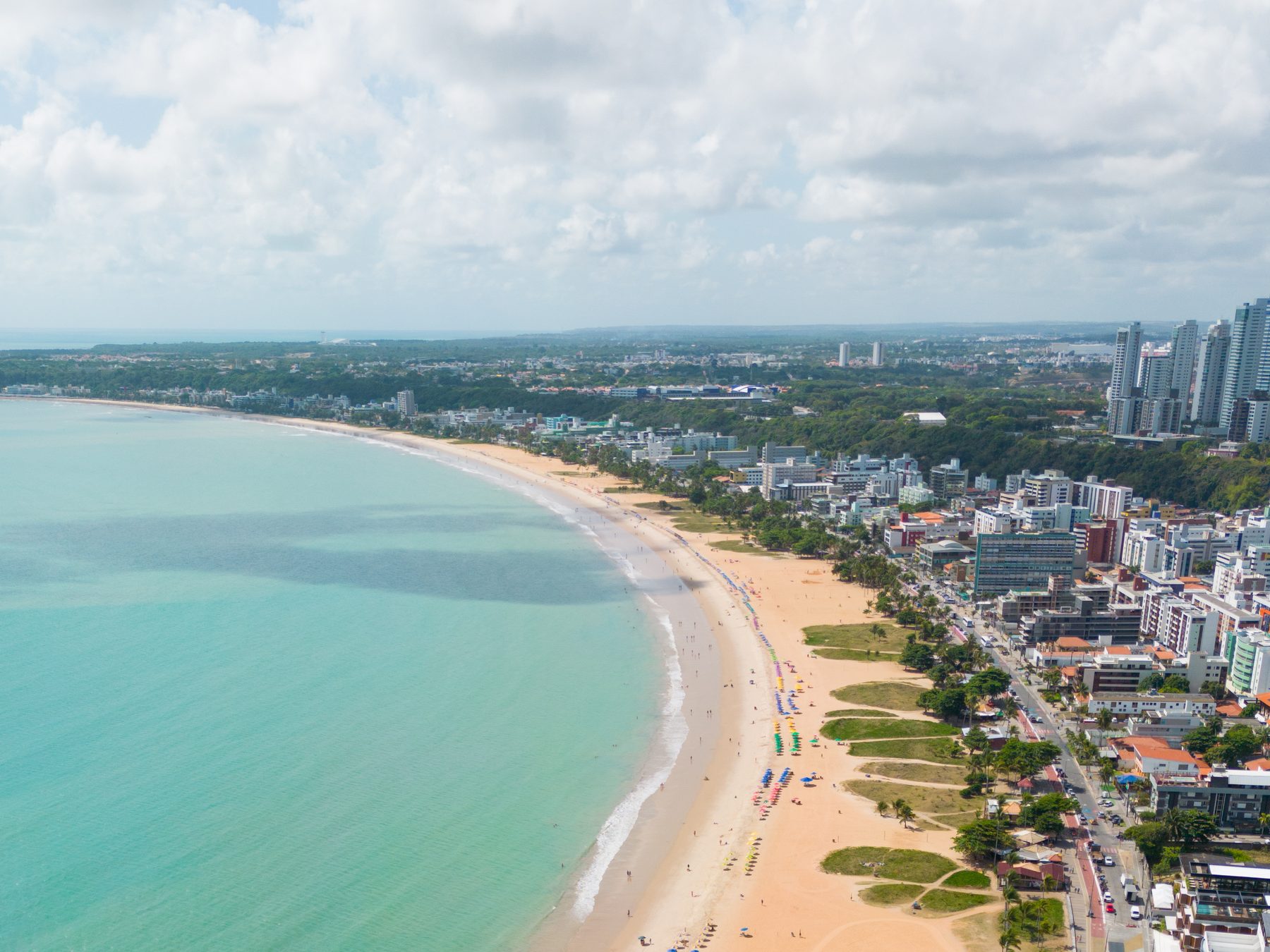 Vista aérea da orla de Cabo Branco em João Pessoa, com o mar verde esmeralda, a faixa larga de areia com barracas coloridas e os prédios da avenida beira mar ao fundo