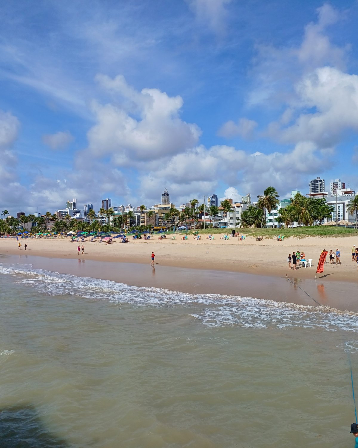 Banhistas na areia e na beira d'água da praia de Tambaú, em João Pessoa, com guarda-sóis coloridos e os prédios da orla ao fundo