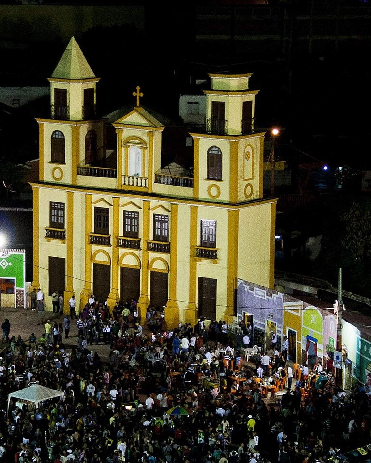 Multidão no Parque do Povo durante o São João de Campina Grande, com a cidade cenográfica iluminada, a igreja amarela e a pirâmide de fogueira acesa