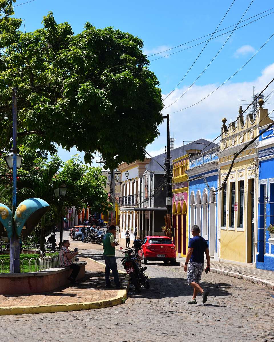 Rua de paralelepípedo no centro histórico de Areia, com casario colonial azul, amarelo e vermelho, praça arborizada e pessoas caminhando