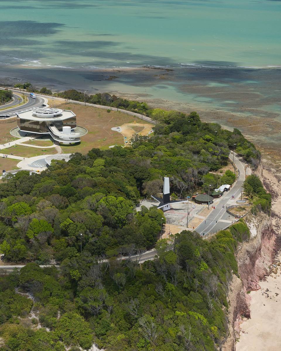 Vista aérea da falésia do Cabo Branco em João Pessoa, com o Farol do Cabo Branco no alto, o mirante, a mata em volta e a praia ao lado do mar esverdeado