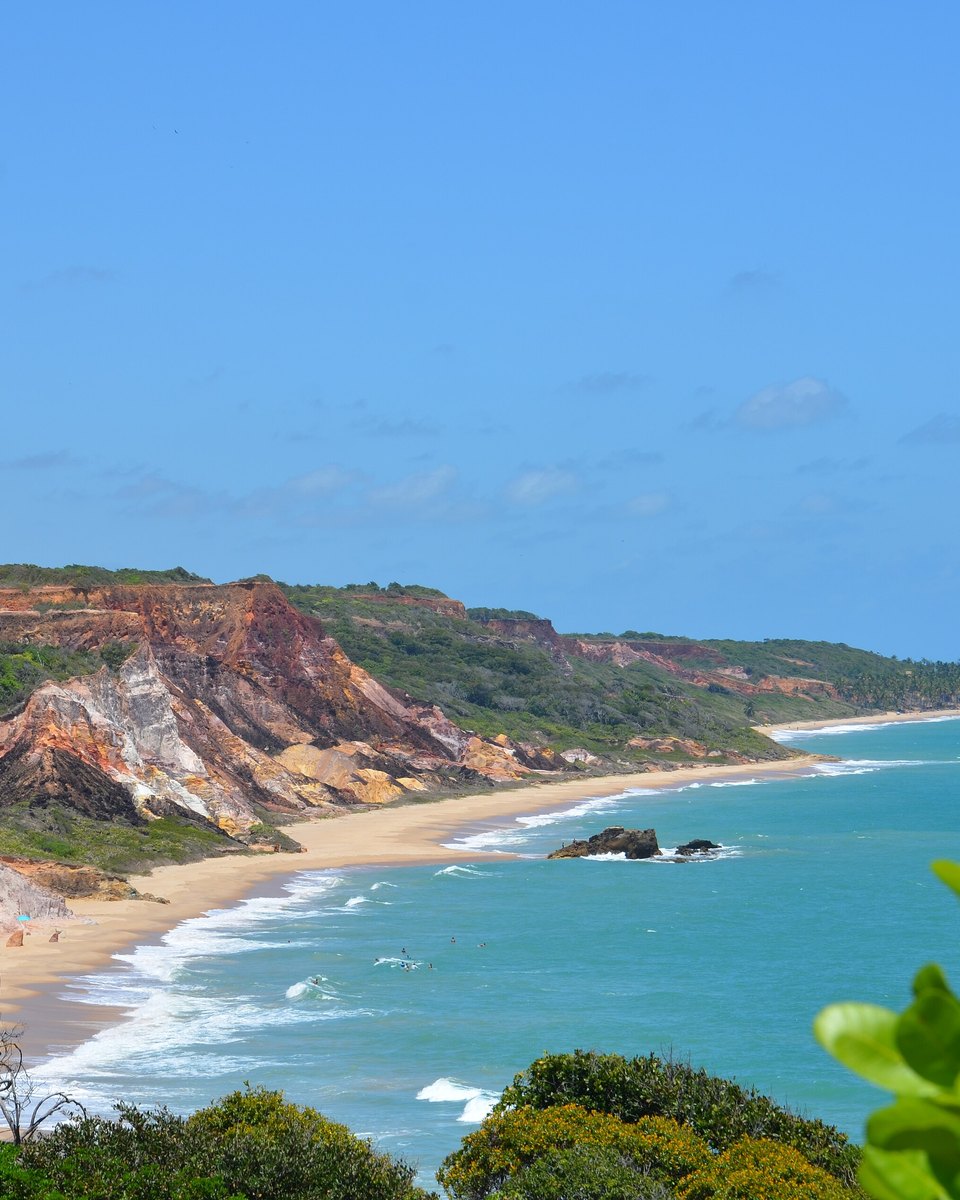 Falésias avermelhadas e faixa de areia da Praia de Coqueirinho, no litoral sul da Paraíba, com mar azul-turquesa e coqueiros ao longe