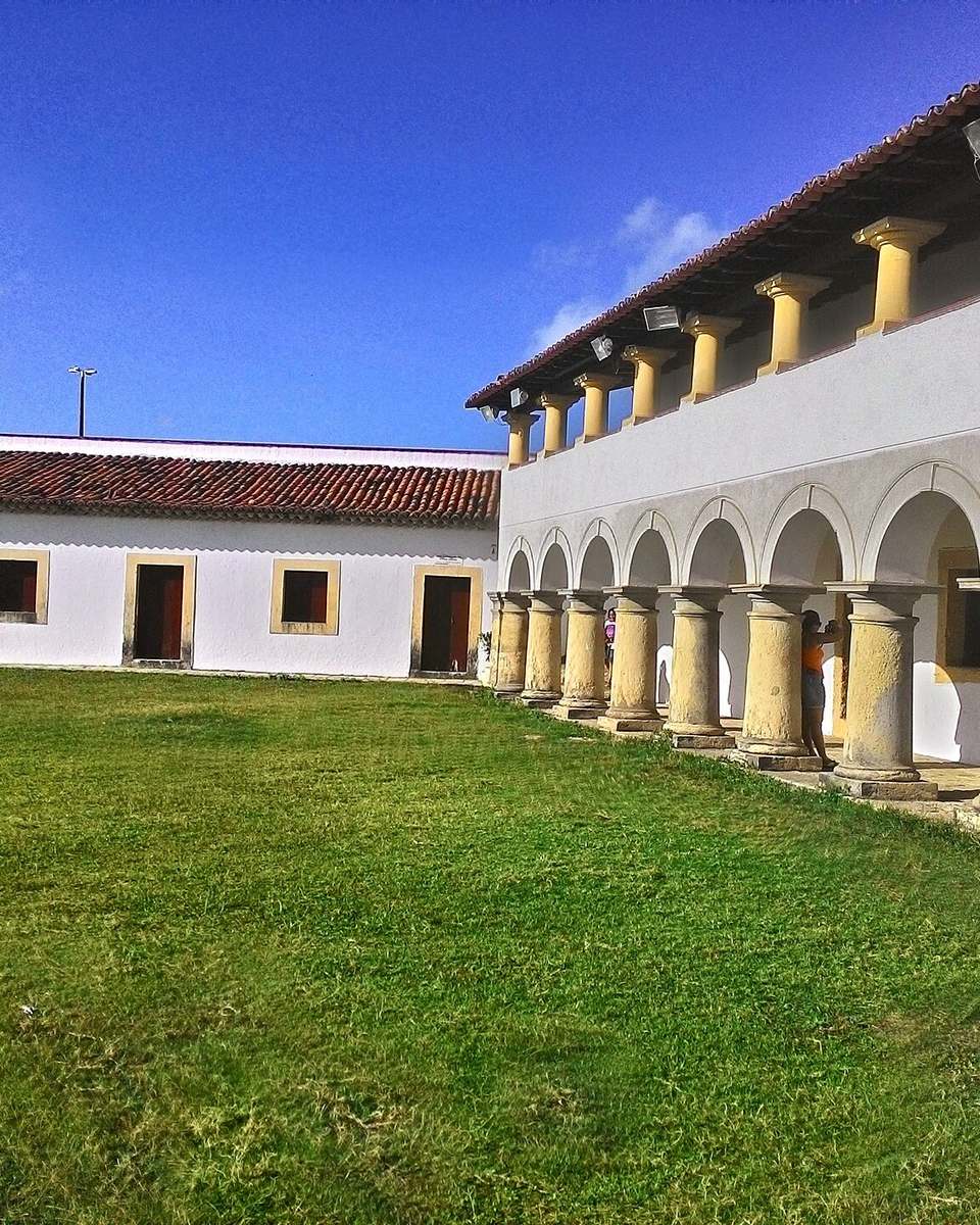 Pátio interno da Fortaleza de Santa Catarina, em Cabedelo, com a arcada de colunas amarelas e o gramado sob o céu azul