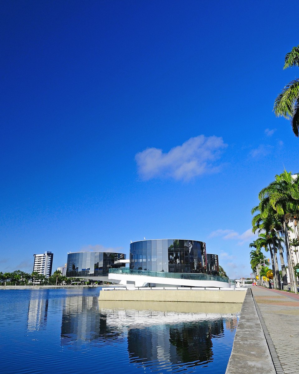 Orla do Açude Velho em Campina Grande, com o prédio circular do Museu de Arte Popular da Paraíba refletido na água e coqueiros ao longo do calçadão