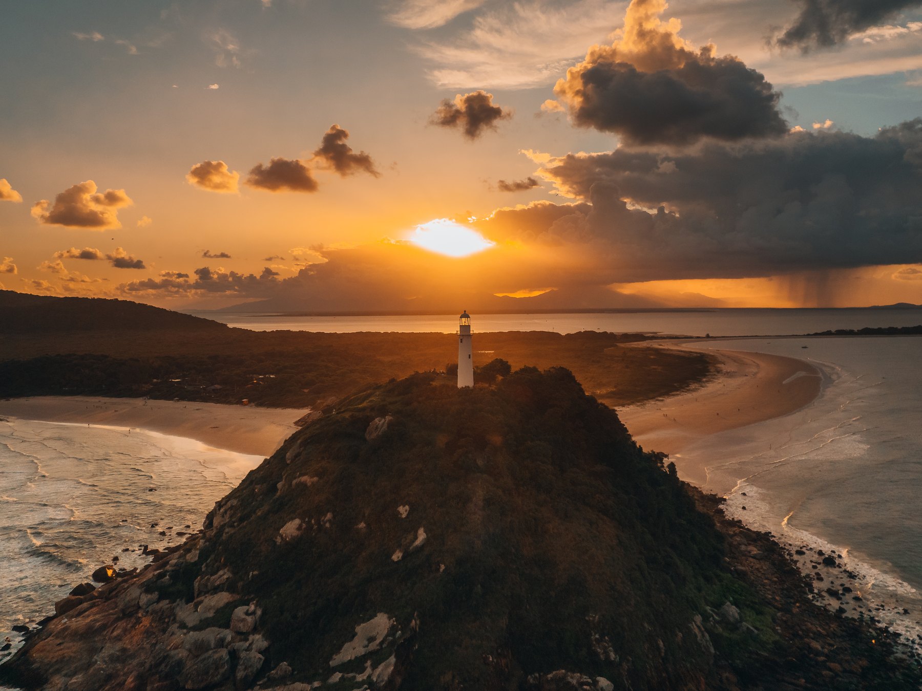 Farol das Conchas no alto de um morro da Ilha do Mel ao pôr do sol, com as praias e o mar dos dois lados e o céu alaranjado.