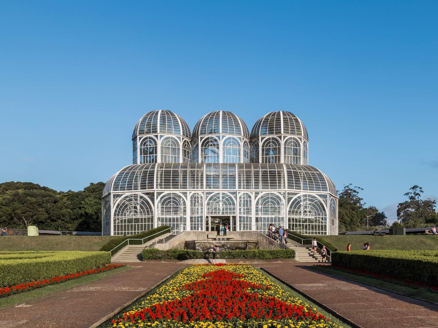 Estufa de vidro e ferro do Jardim Botânico de Curitiba, com canteiros de flores vermelhas e amarelas no jardim à frente e céu azul.