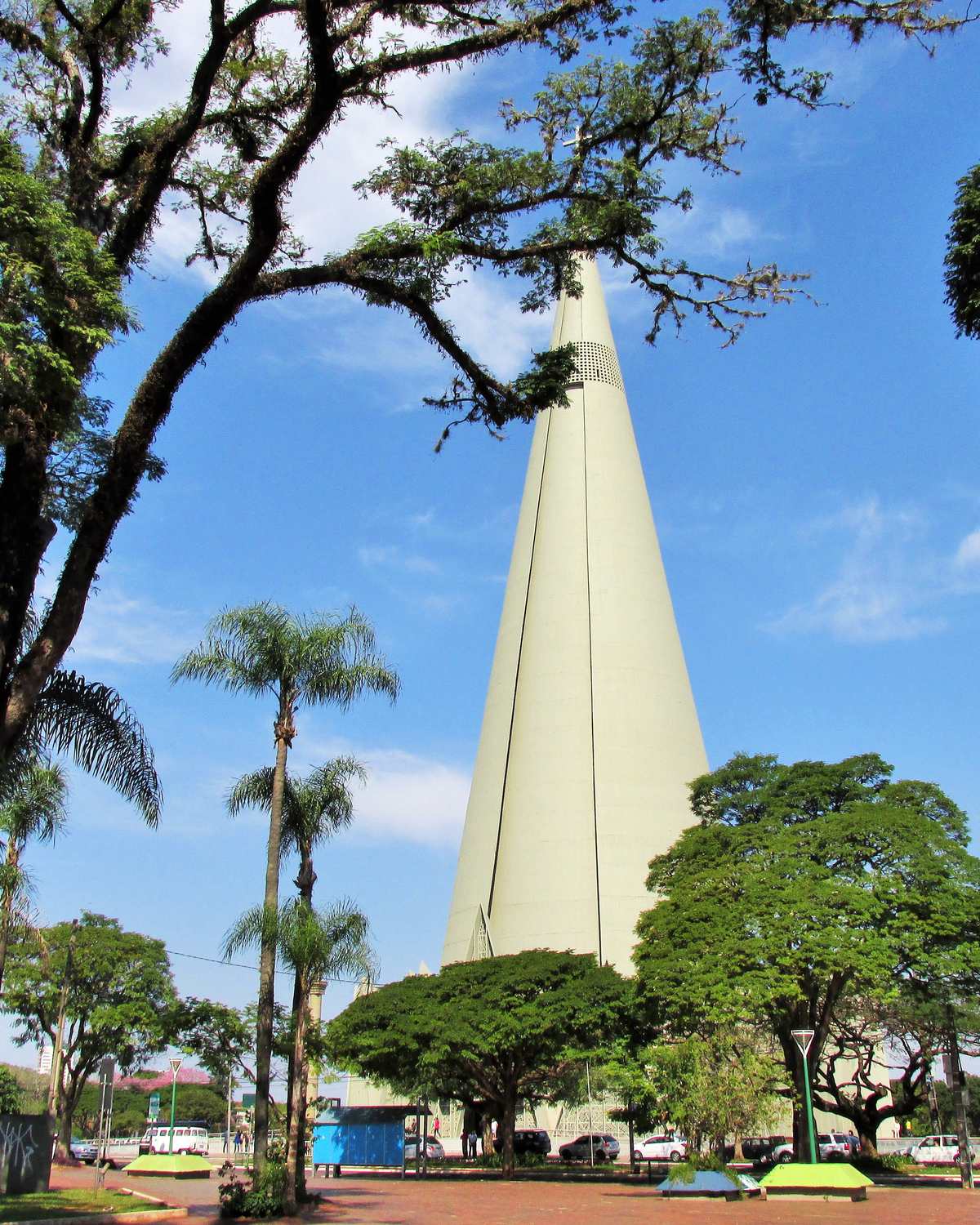 Torre cônica da Catedral de Maringá vista da praça, entre árvores e palmeiras, num dia de céu azul.