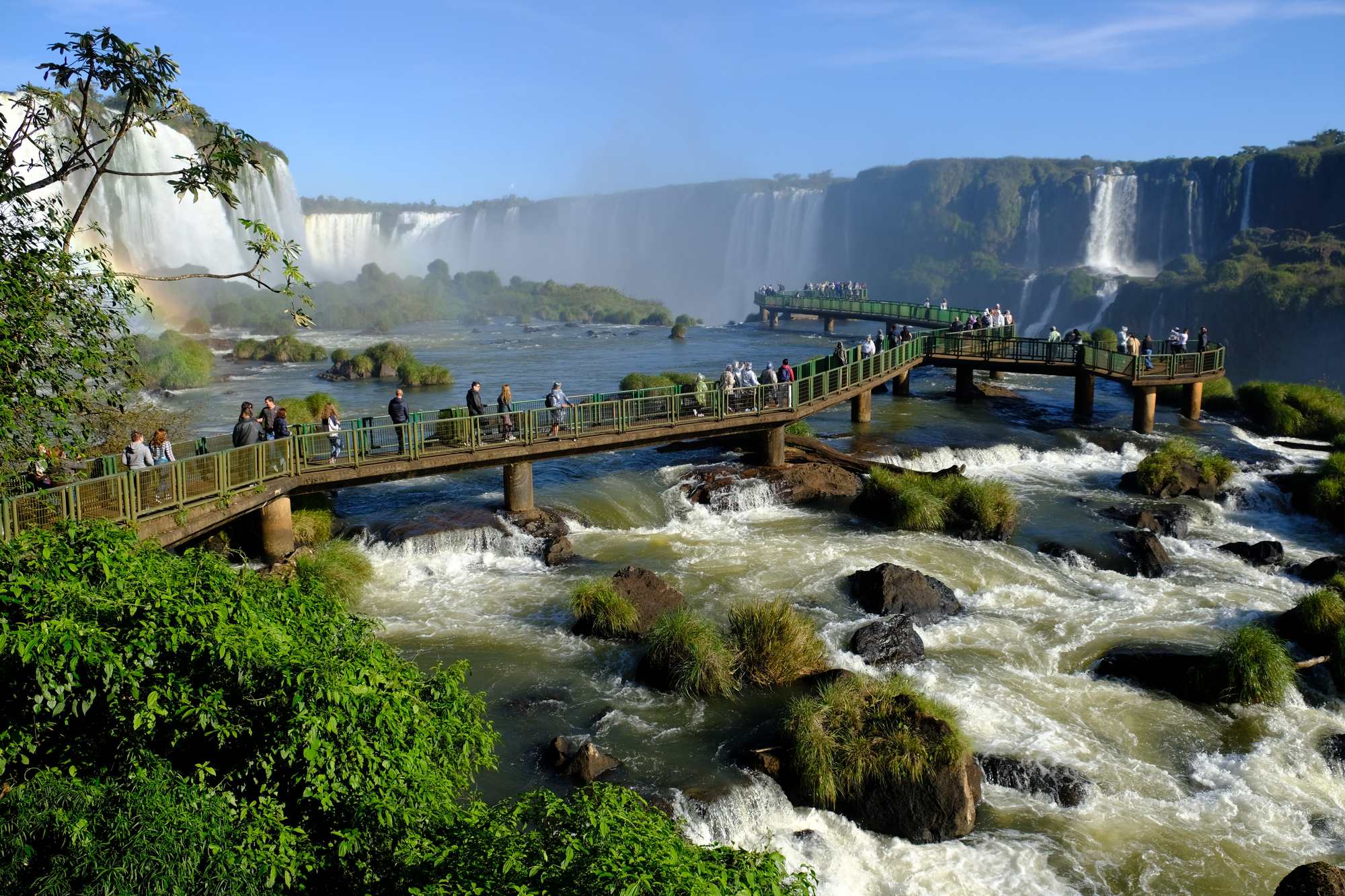 Turistas caminhando na passarela sobre as corredeiras do rio Iguaçu, com as quedas das Cataratas ao fundo, no lado brasileiro, em Foz do Iguaçu, no Paraná.