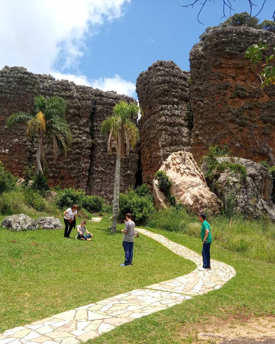 Visitantes caminhando numa trilha de pedra entre o gramado e os grandes blocos de arenito avermelhado do Parque Estadual de Vila Velha, em Ponta Grossa.