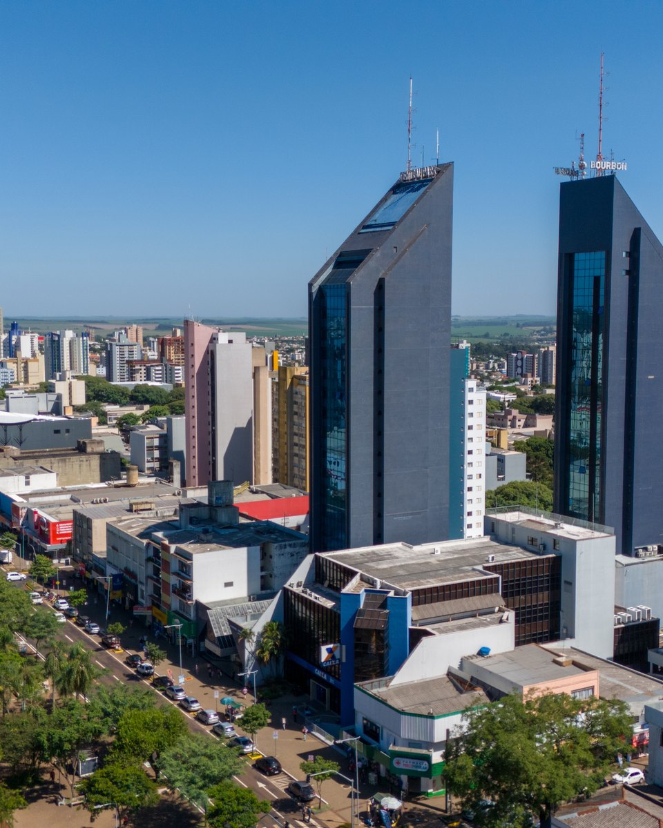 Vista aérea do centro de Cascavel, com a Avenida Brasil arborizada, lojas no térreo, duas torres espelhadas e prédios residenciais até o horizonte, num dia de céu azul.