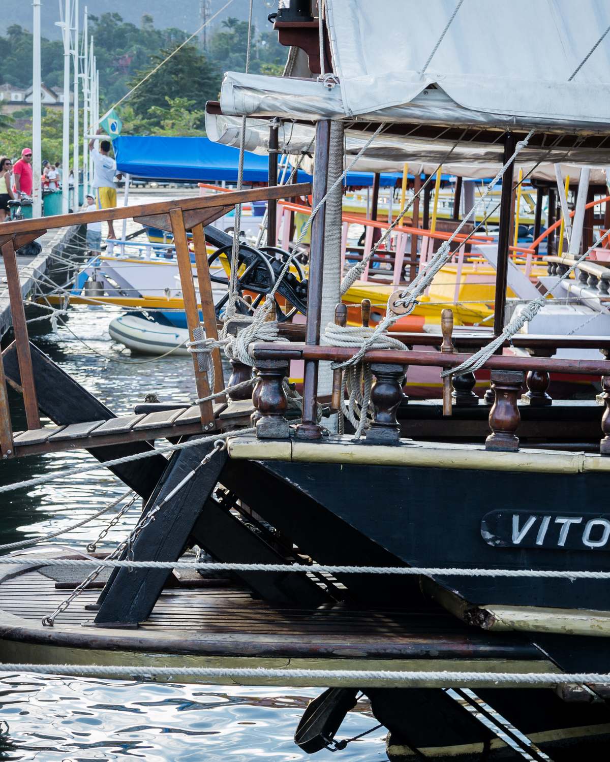 Escuna de madeira atracada no cais de Paraty, com turistas caminhando pelo píer e outras escunas coloridas ao fundo.