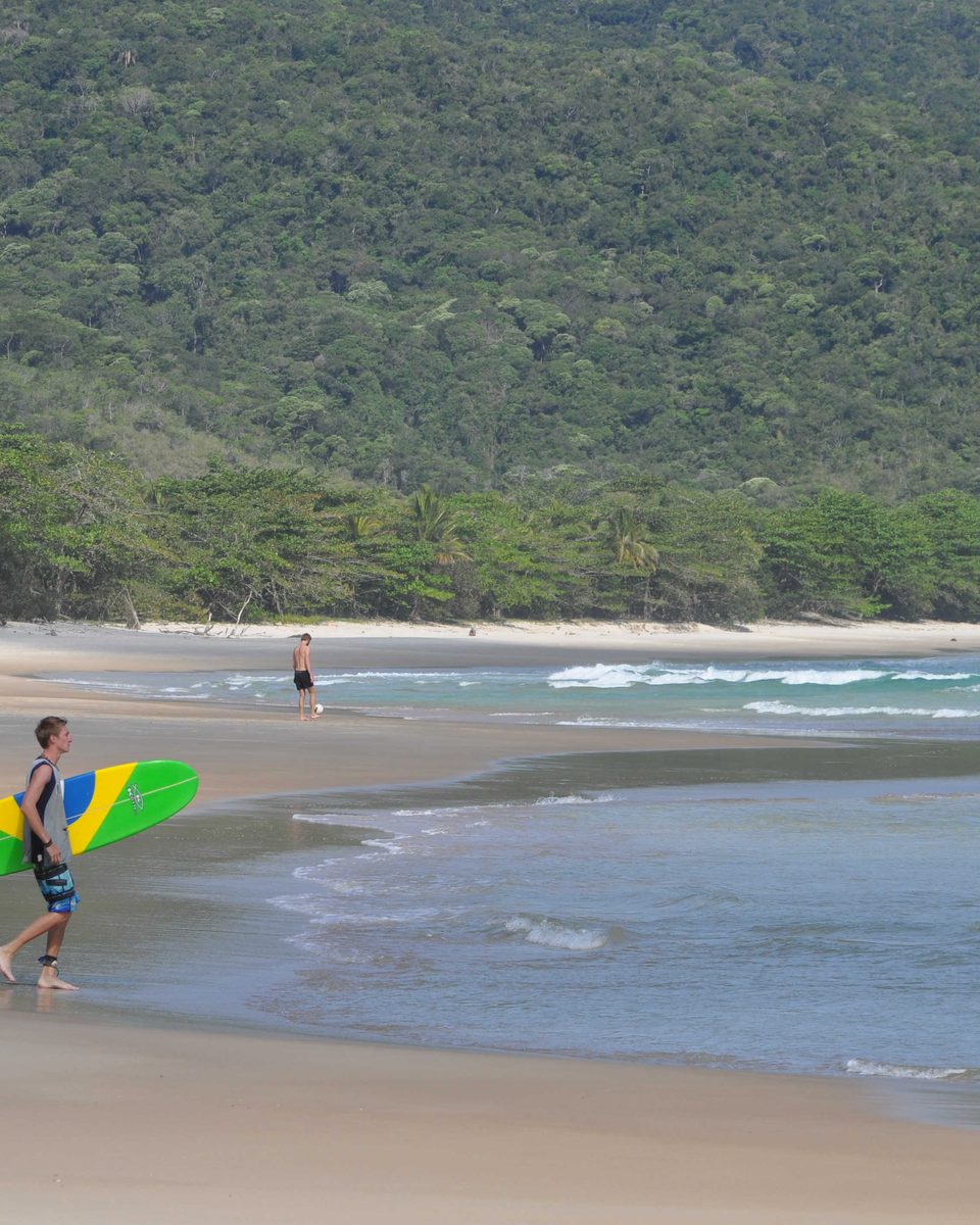 Surfista com prancha na beira da praia, com a mata da Costa Verde ao fundo.