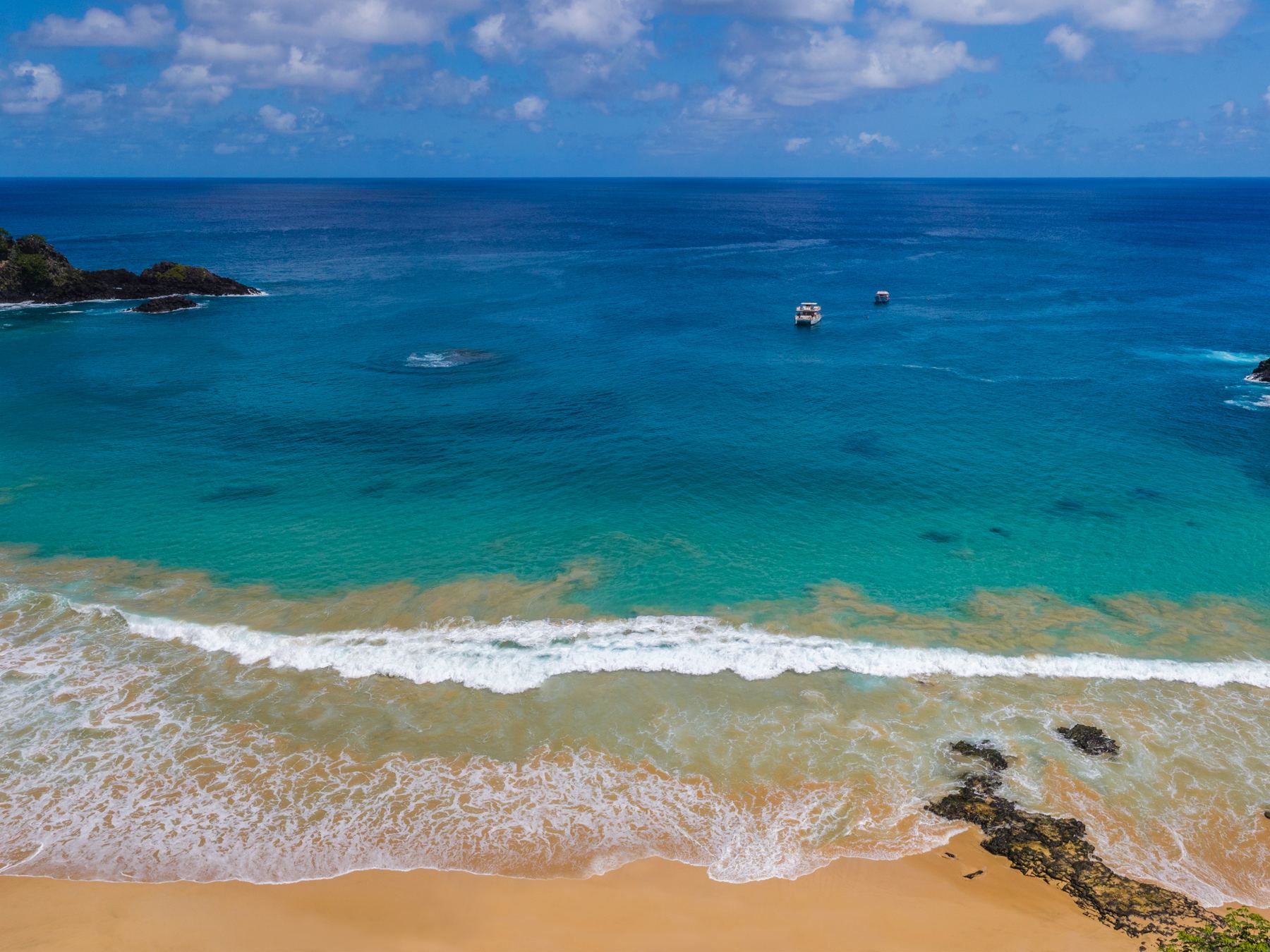 Panorama da Baía do Sancho, em Fernando de Noronha, com a faixa de areia dourada entre falésias cobertas de mata, ondas pequenas e o mar azul-turquesa até o horizonte.