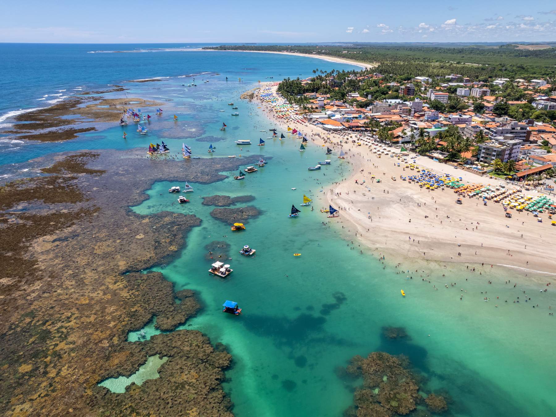 Vista aérea das piscinas naturais de Porto de Galinhas, com os recifes escuros na água verde-clara, jangadas, banhistas e a praia cheia de guarda-sóis coloridos.