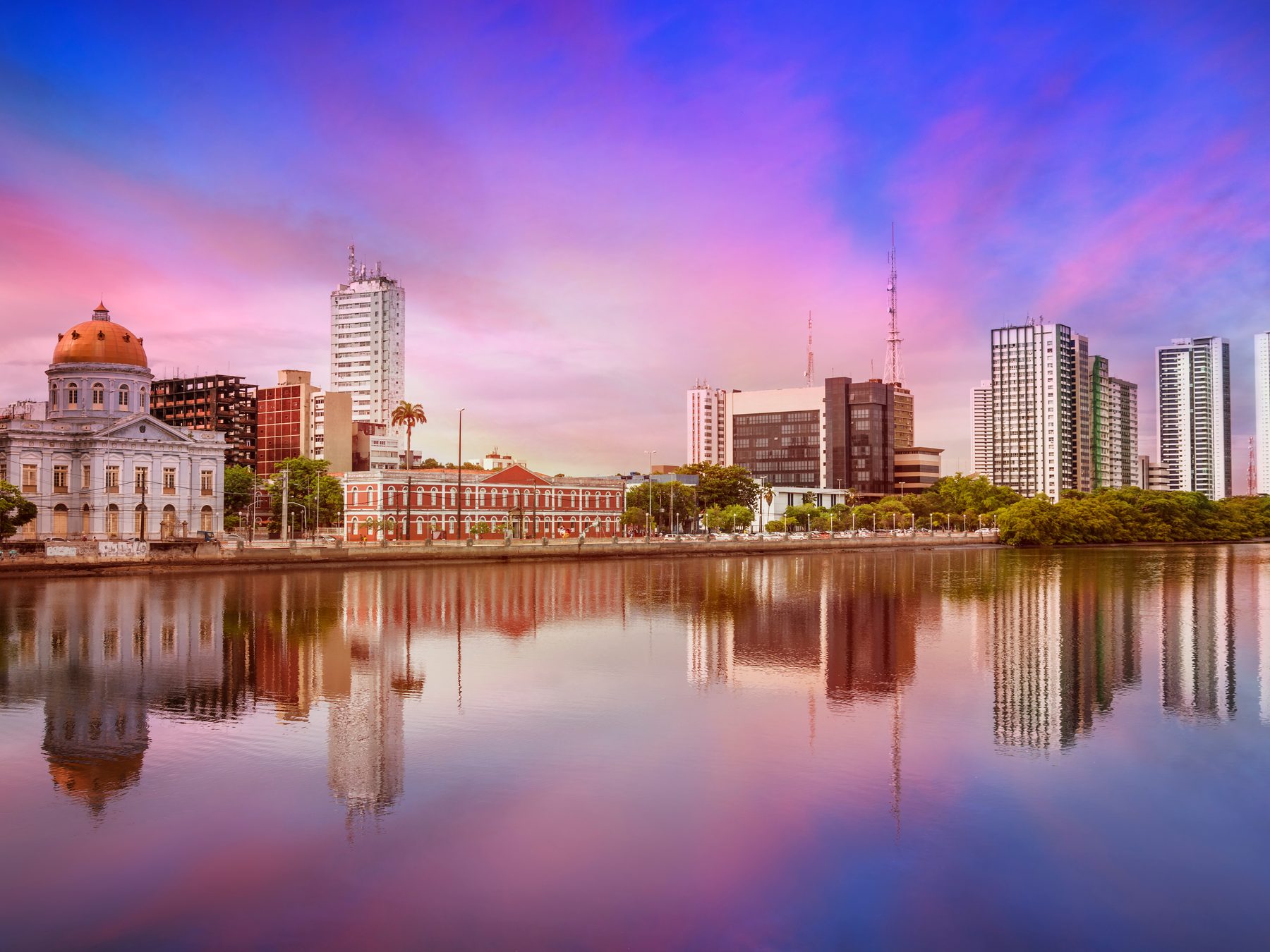 Casarões e prédios da margem do Rio Capibaribe refletidos na água parada ao entardecer, com o céu rosa e lilás sobre o centro do Recife.