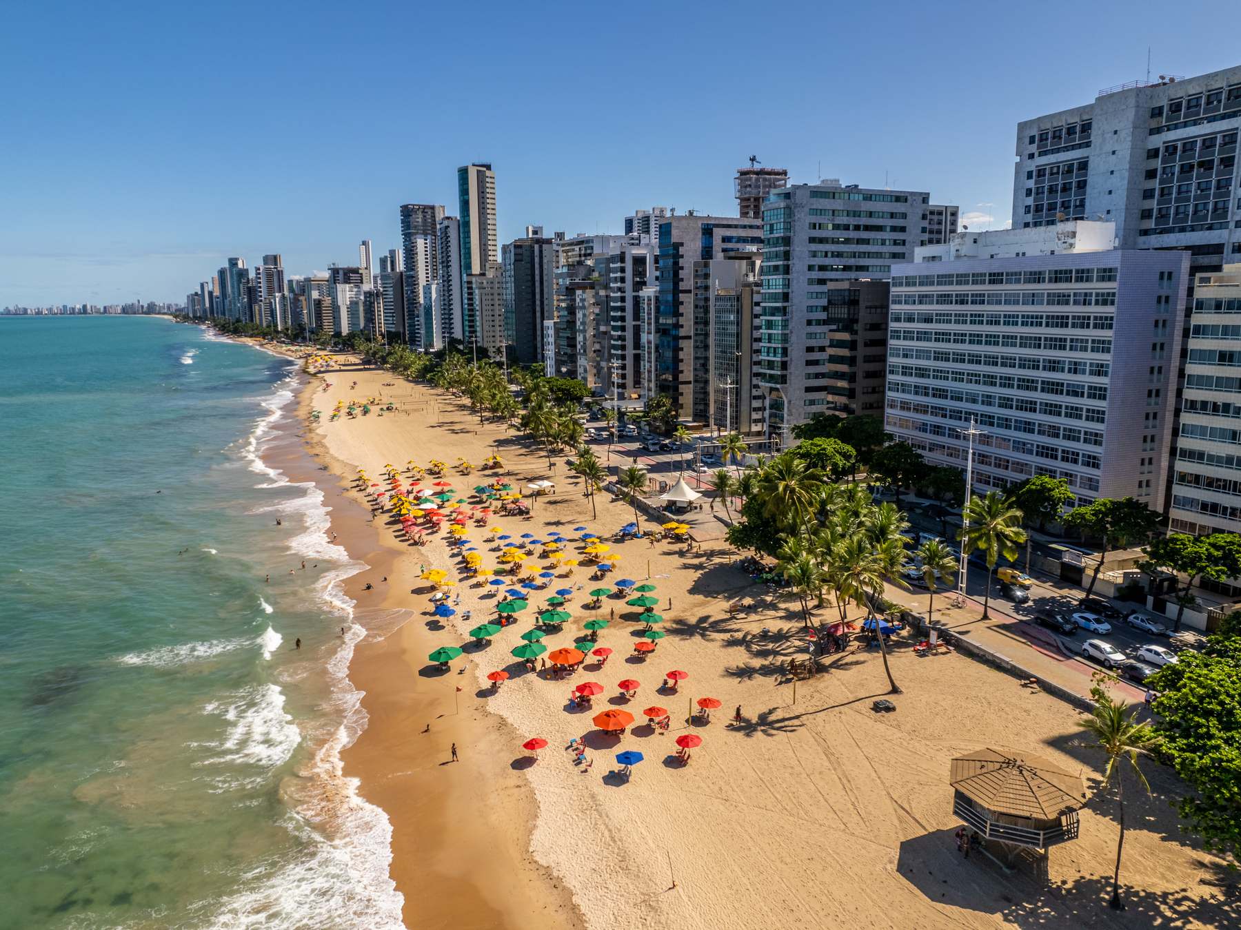 Vista aérea da Praia de Boa Viagem, com guarda-sóis coloridos na areia, o mar esverdeado e a fileira de prédios da orla até o horizonte.