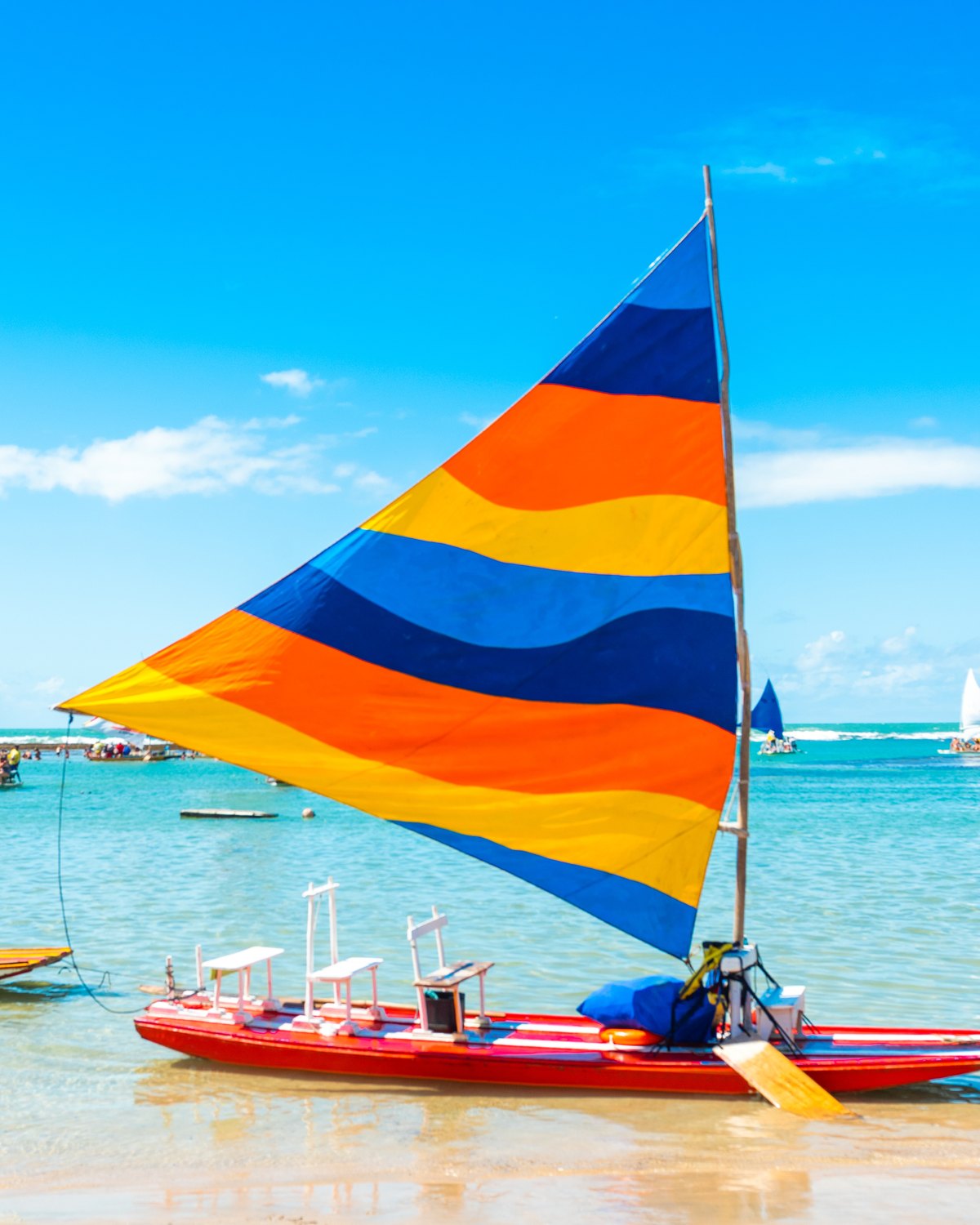 Jangada de vela listrada em laranja, azul e amarelo parada na areia molhada de Porto de Galinhas, com o mar azul-turquesa e outras jangadas ao fundo.
