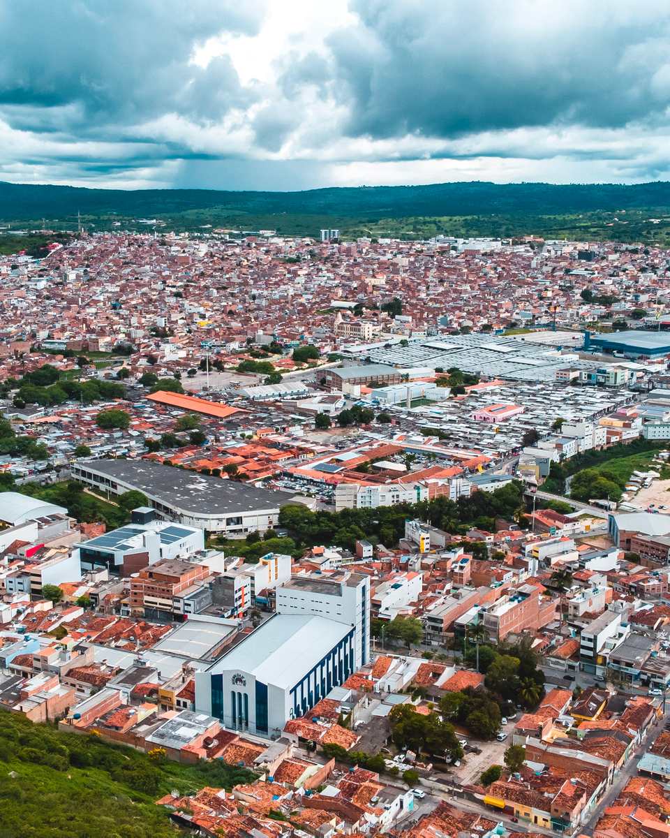 Vista aérea de Caruaru, com o casario de telhados vermelhos, avenidas e prédios se espalhando até a serra no horizonte, sob céu com nuvens.
