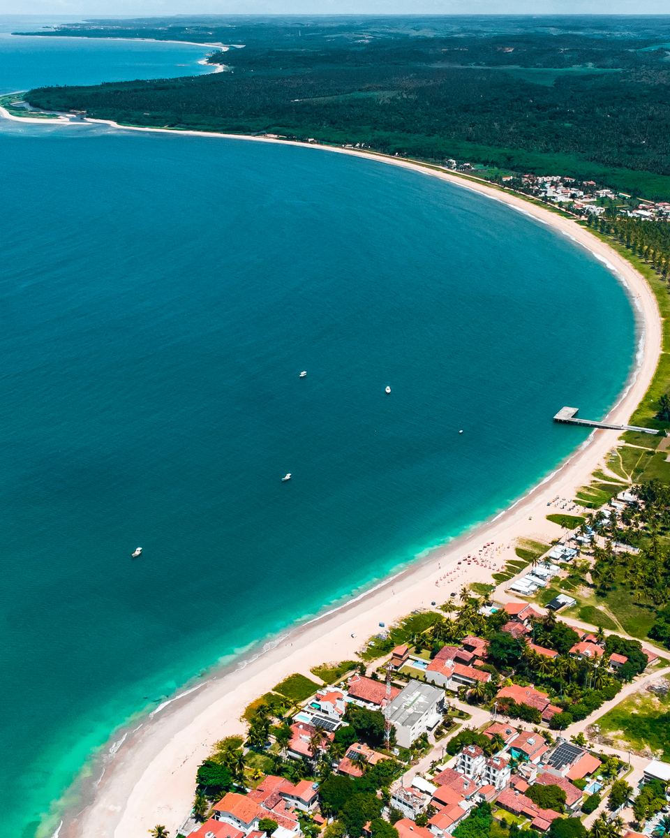 Vista aérea da baía de Tamandaré, com o mar azul-turquesa e verde, a longa faixa de areia branca em curva e as casas entre coqueiros e mata.