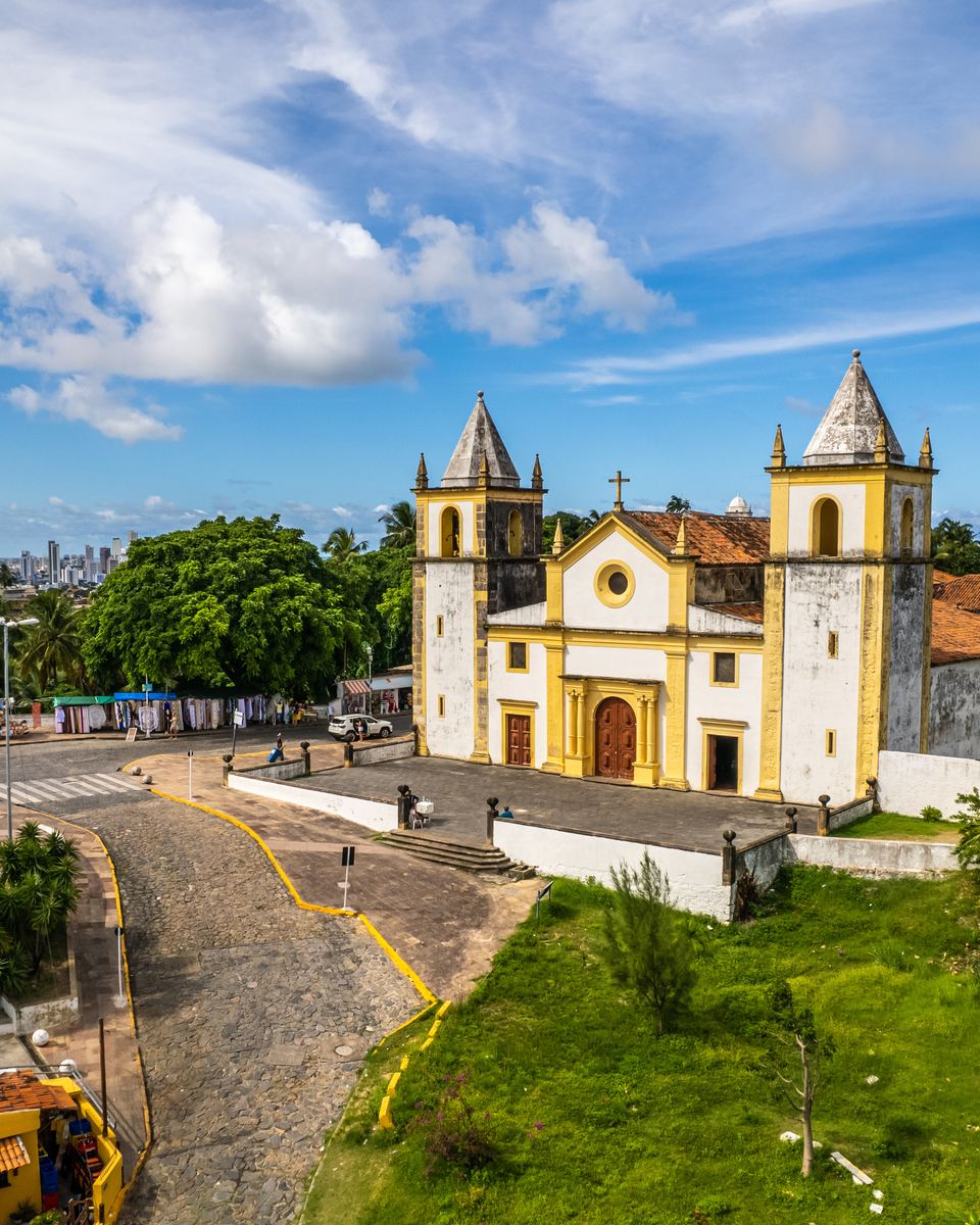 Alto da Sé, em Olinda, visto do alto, com a igreja da Sé de duas torres, coqueiros, a ladeira de pedra com pessoas caminhando e o mar ao fundo.