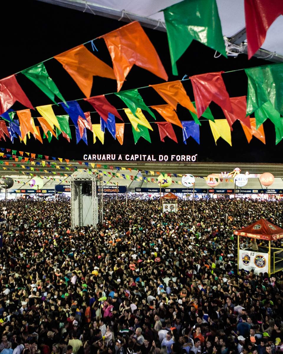 Multidão no Pátio do Forró à noite durante o São João de Caruaru, sob bandeirinhas coloridas, com o palco iluminado e a faixa Caruaru a capital do forró ao fundo.