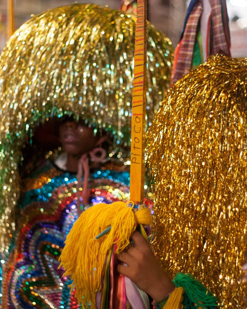 Caboclo de lança do maracatu rural com cabeleira de fitas douradas e brilhantes e gola bordada de lantejoulas coloridas, segurando a lança enfeitada com fitas, entre outros brincantes numa apresentação no Recife.