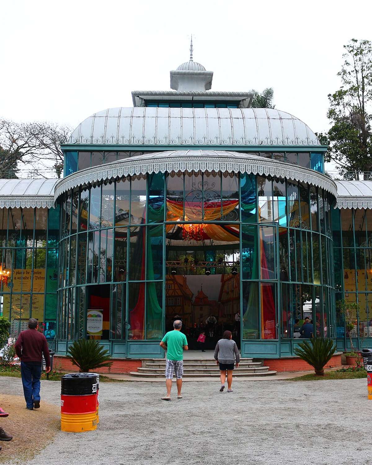Visitantes passeando e sentados nos bancos diante do Palácio de Cristal, em Petrópolis, com o pavilhão de ferro e vidro decorado com tecidos coloridos.