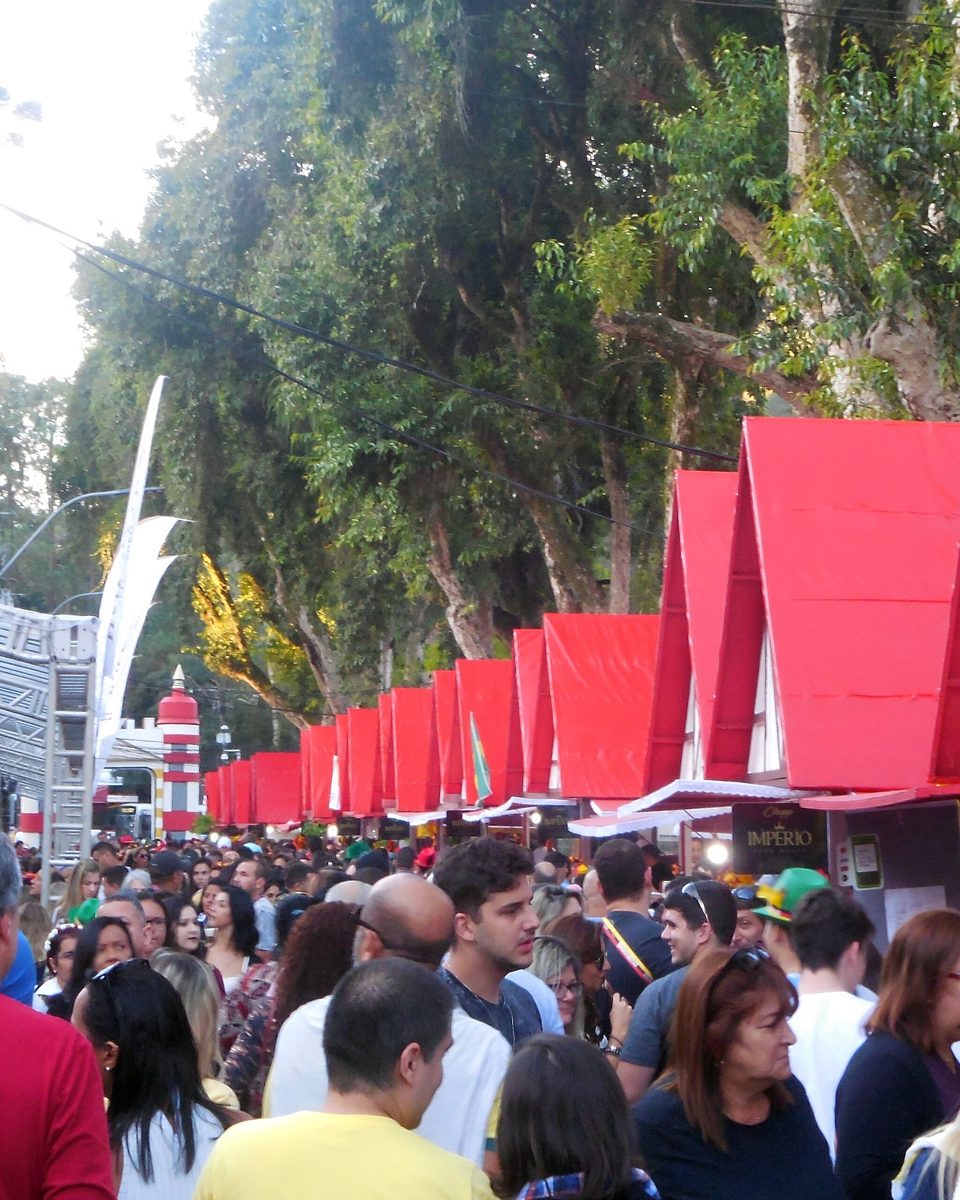 Multidão de adultos caminhando entre as barracas de telhado vermelho em forma de chalé na Bauernfest, em Petrópolis, com árvores altas e o palco ao fundo.
