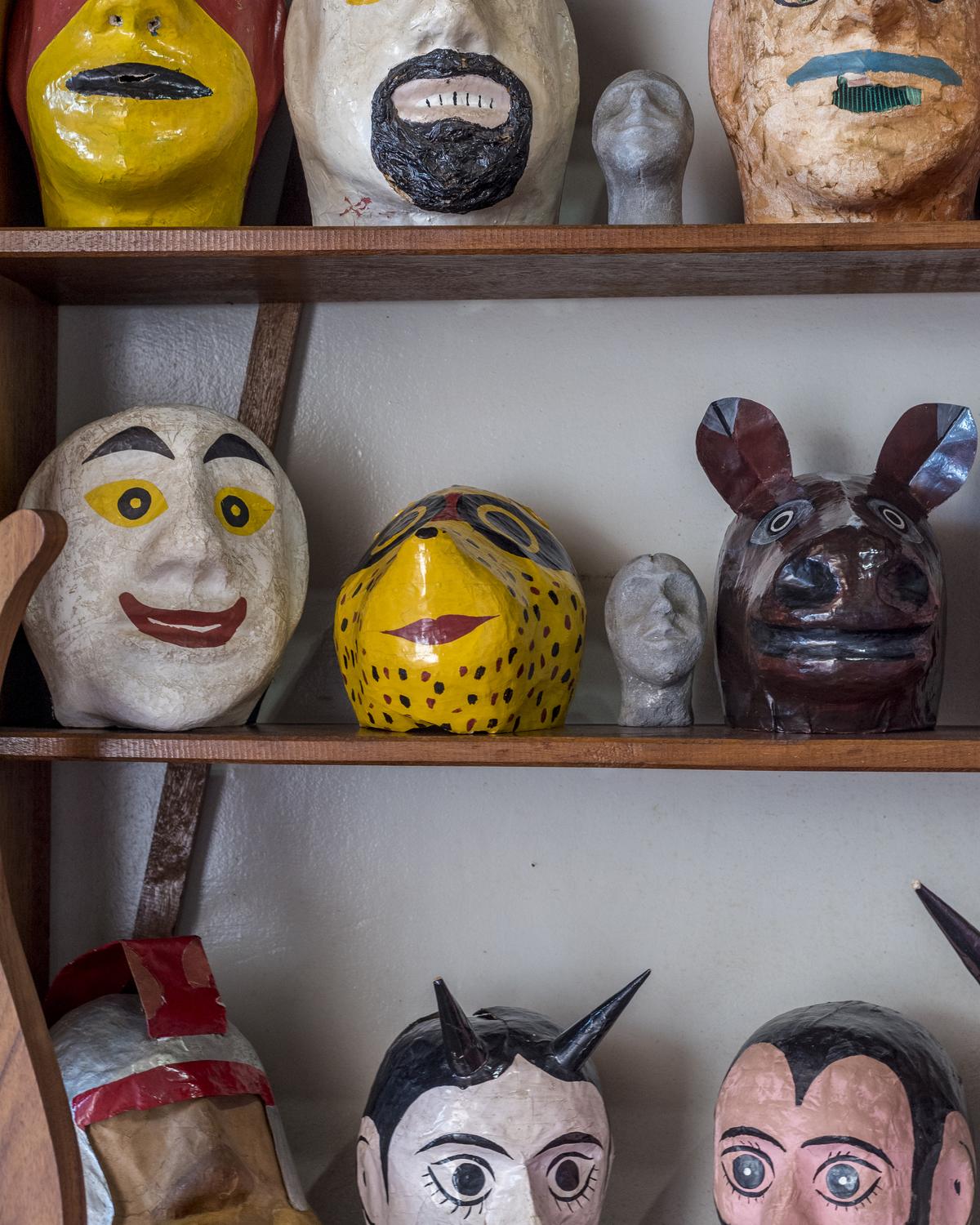 Máscaras de mascarados das Cavalhadas de Pirenópolis, de papel machê pintado, com bigodes, chifres e bocas abertas, enfileiradas em prateleiras de madeira numa casa do centro histórico.