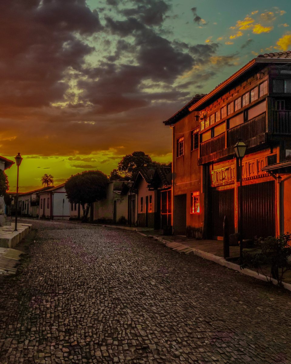 Rua da Prata, no centro histórico de Pirenópolis, ao pôr do sol, com o calçamento de pedra irregular, casarões coloniais de sacada de madeira dos dois lados e o céu laranja e rosa com nuvens.