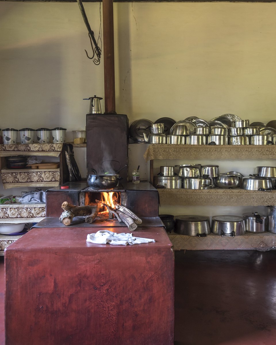 Cozinha antiga da Fazenda Babilônia, em Pirenópolis, com fogão a lenha aceso, bancada de alvenaria vermelha, prateleiras cheias de panelas de alumínio e travessas, e uma janela de madeira aberta.