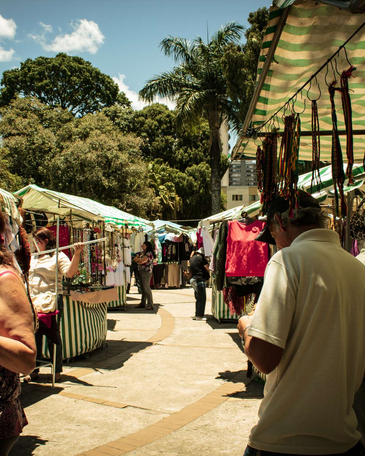 Mulher escolhendo uma blusa pendurada numa barraca de feira de rua no centro de Poços de Caldas, com toldos listrados de verde e branco e outros clientes olhando colares e roupas.