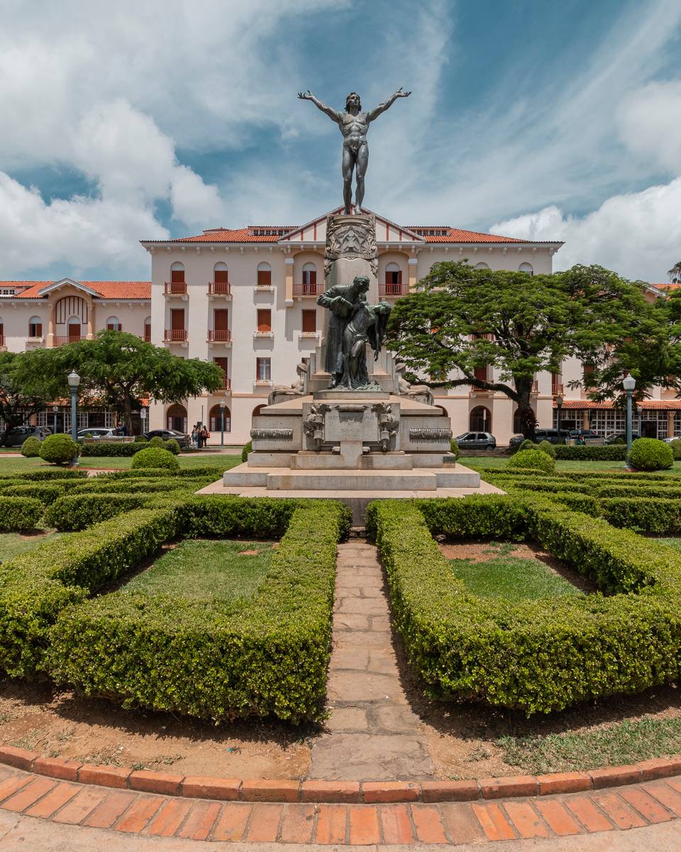 Fachada do Palace Hotel de Poços de Caldas, com telhado de telhas vermelhas e janelas brancas, vista da Praça Pedro Sanches, com o monumento de bronze no centro e canteiros de buxinho em volta.