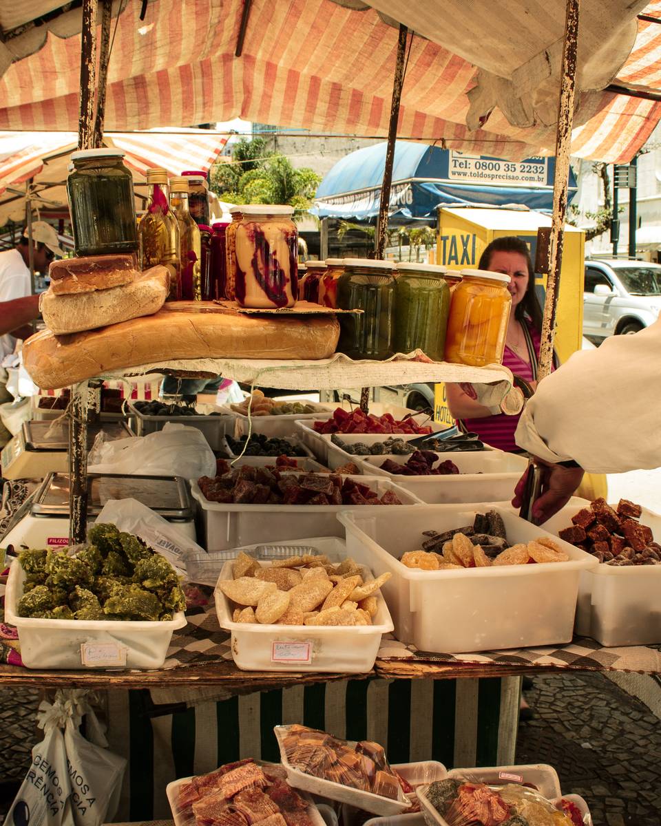 Feirante de camisa clara arrumando uma banca de feira de rua em Poços de Caldas, com queijos, pães, potes de compota e bandejas de quitandas sob um toldo listrado.