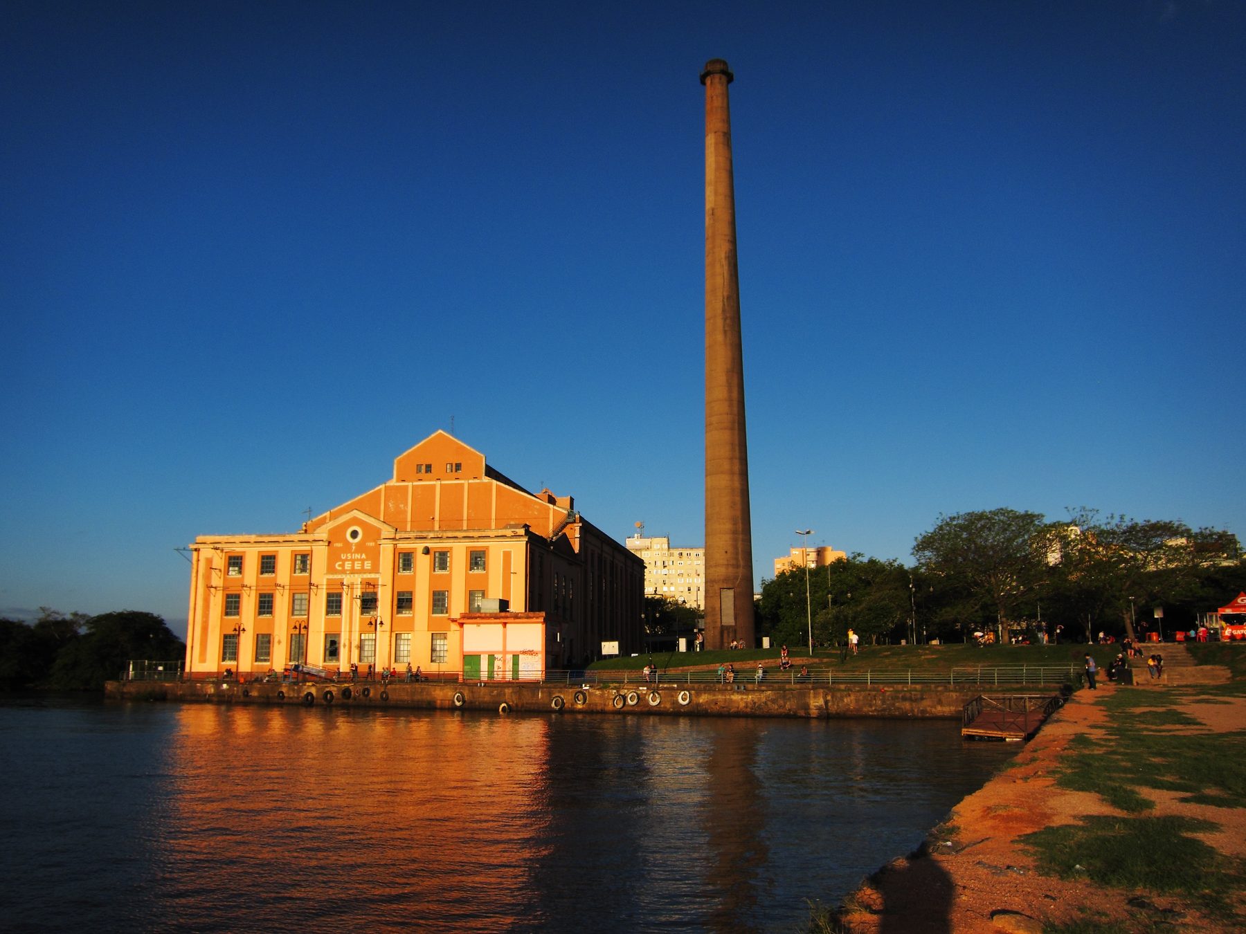 Usina do Gasômetro dourada pelo sol do fim da tarde, com a chaminé alta contra o céu azul-escuro e o prédio refletido na água do Guaíba, em Porto Alegre.