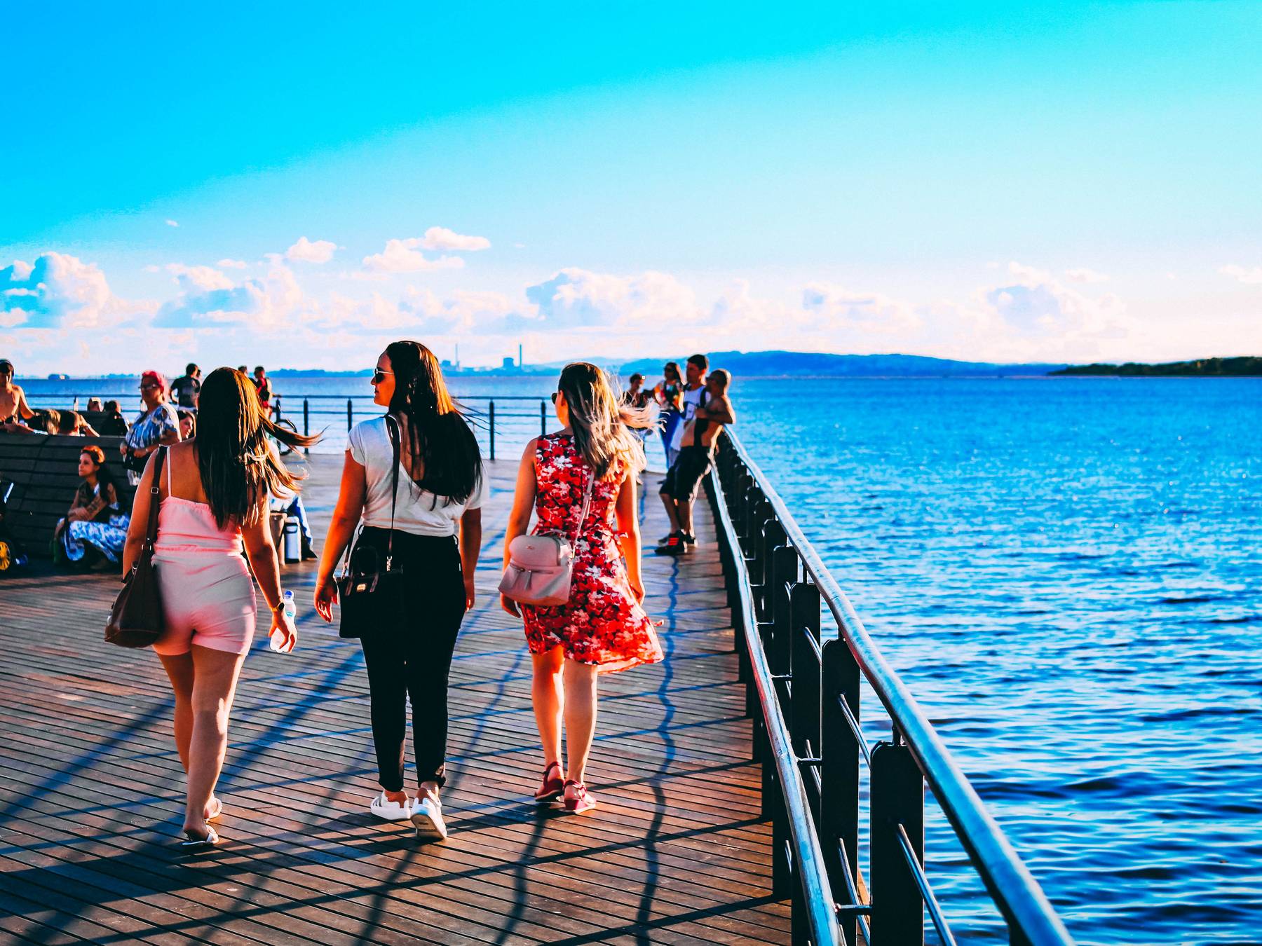 Três amigas caminhando de costas pelo deque de madeira da Orla Moacyr Scliar, em Porto Alegre, ao lado da grade sobre o Guaíba azul, com gente sentada ao fundo num dia de céu claro.