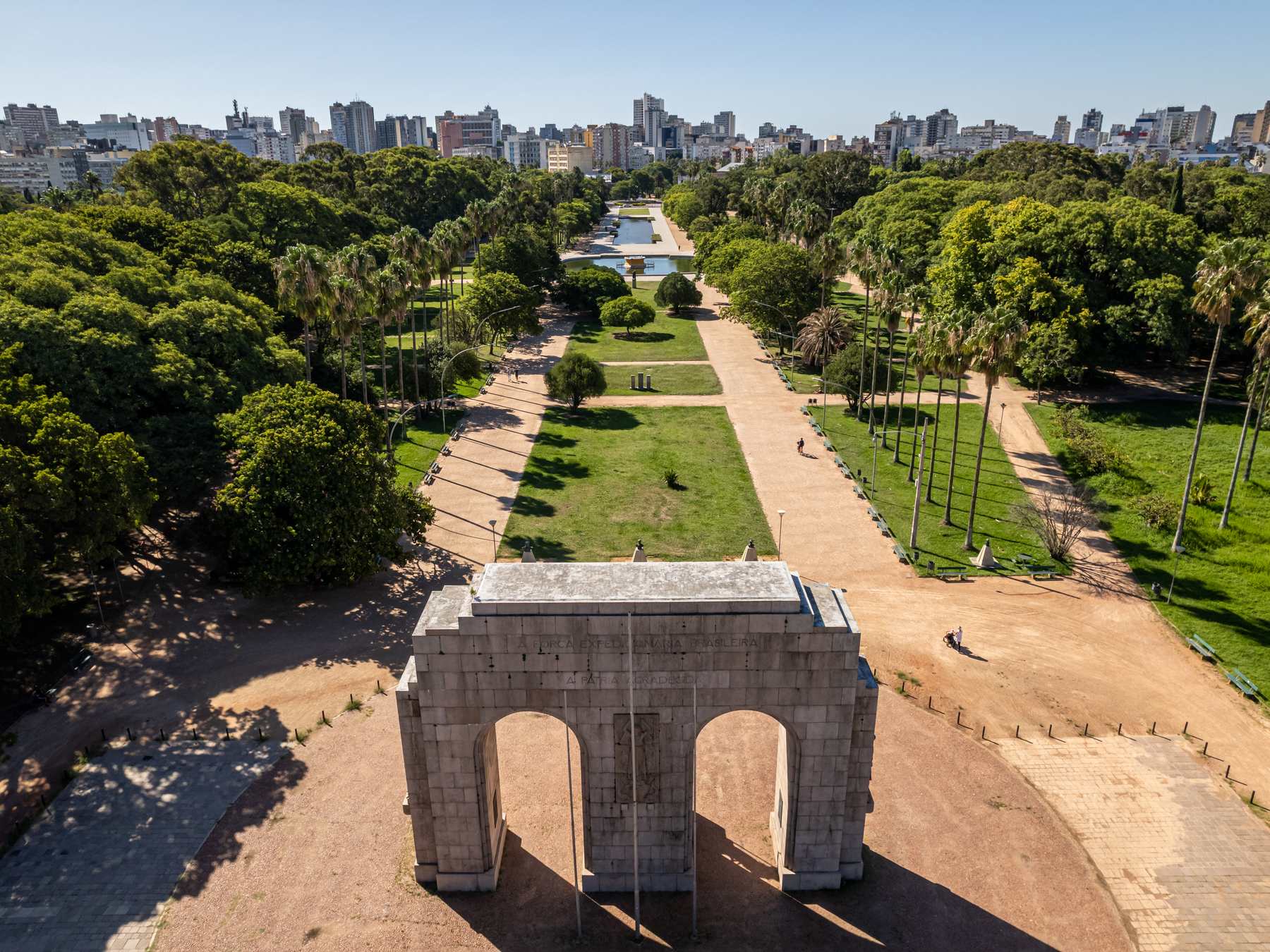 Vista aérea do Parque Farroupilha, a Redenção, em Porto Alegre, com o Monumento ao Expedicionário em primeiro plano, a alameda central entre as árvores e os prédios da cidade no horizonte.