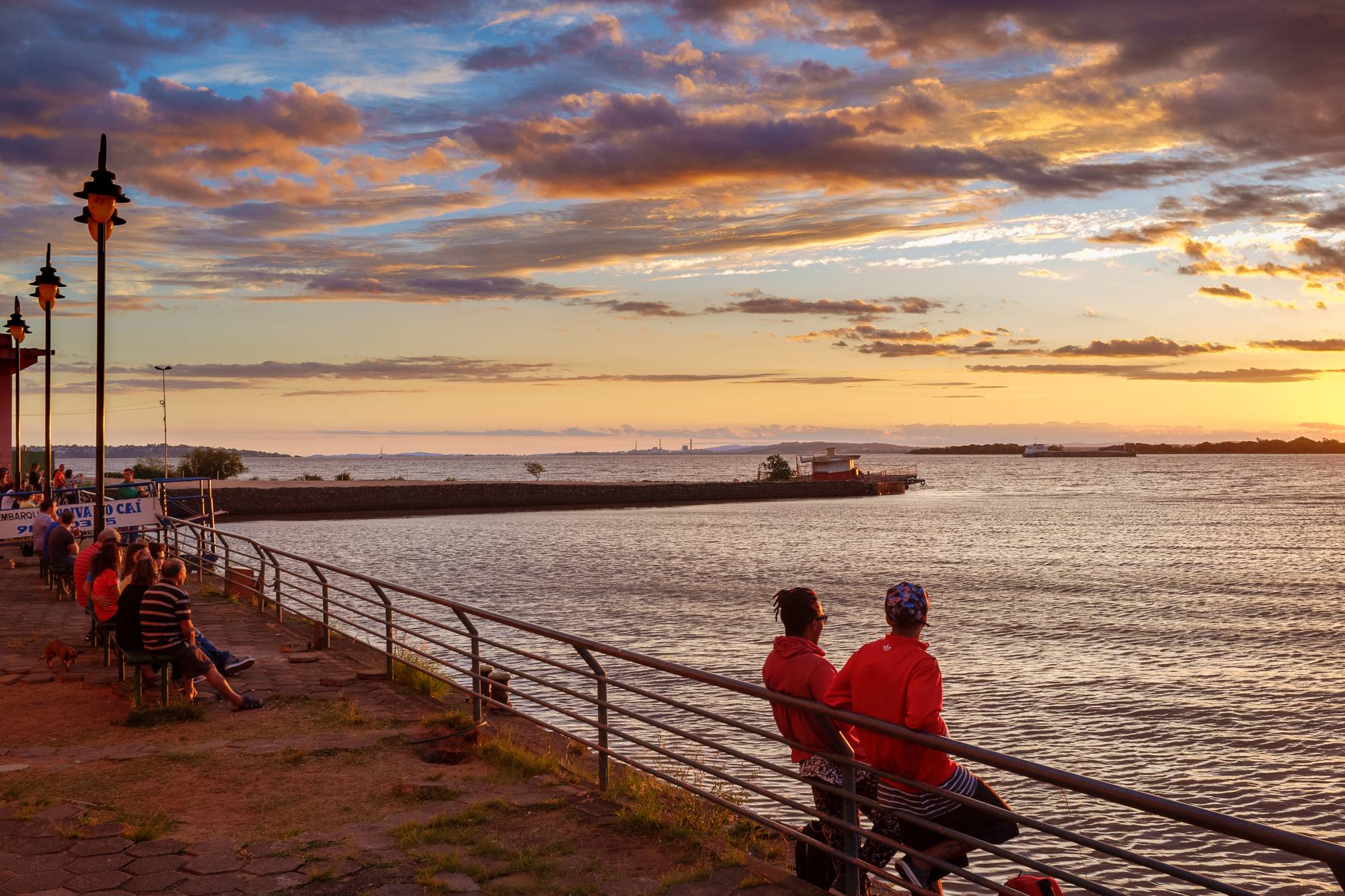 Casal debruçado na grade da orla do Guaíba, perto da Usina do Gasômetro, em Porto Alegre, olhando o pôr do sol sobre a água, com outras pessoas sentadas no calçadão e o céu alaranjado cheio de nuvens.