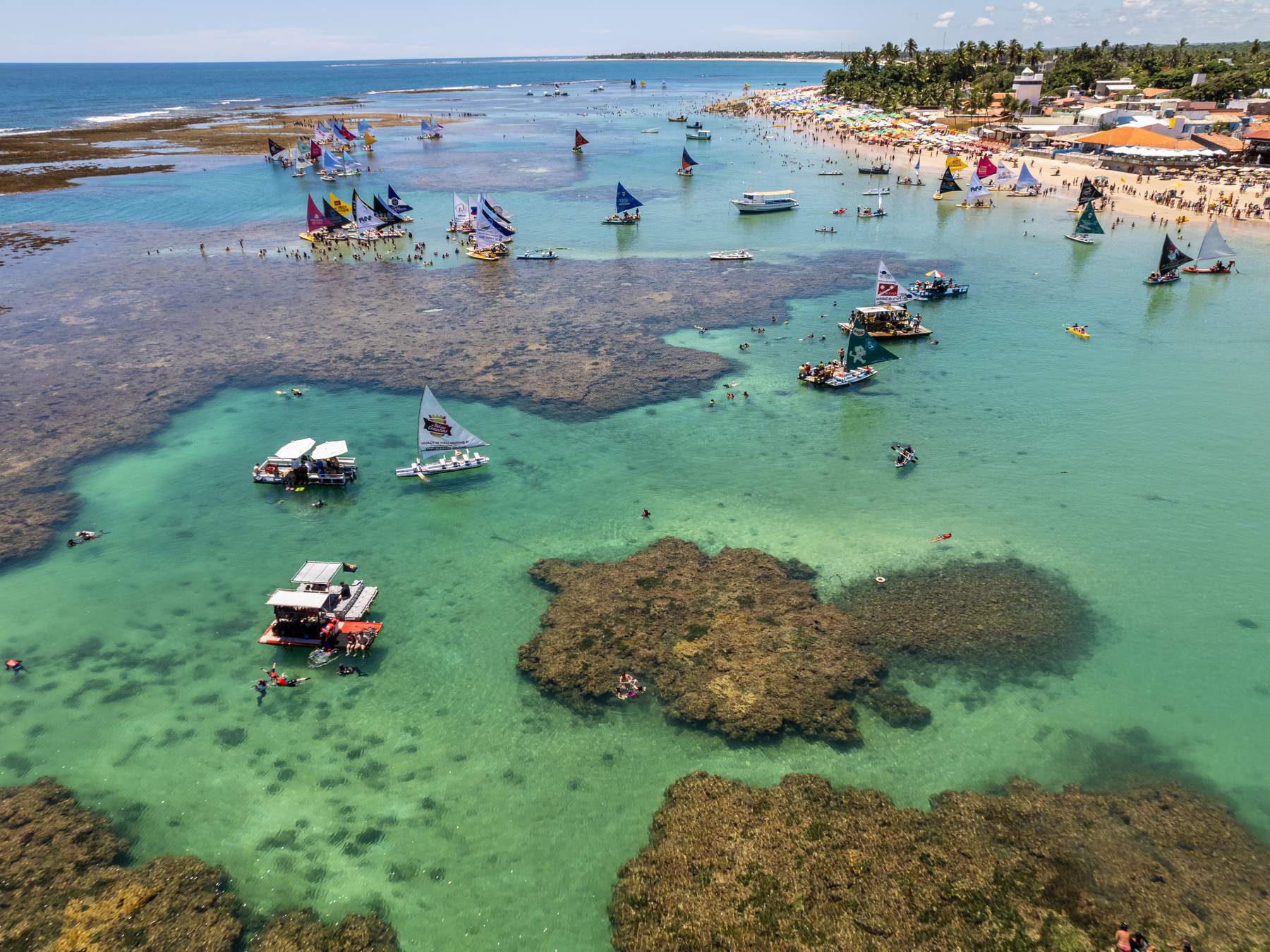 Piscinas naturais de Porto de Galinhas vistas do alto, com os recifes marrons cercando poças de água verde-clara, jangadas de velas coloridas e banhistas na água rasa e a praia com guarda-sóis ao fundo.