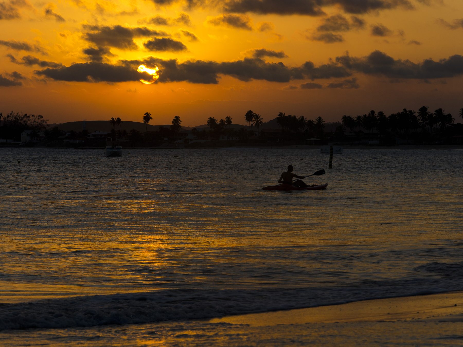 Pôr do sol na Praia de Serrambi, com o céu laranja e nuvens escuras, o reflexo dourado na água calma e a silhueta de uma pessoa remando um caiaque.