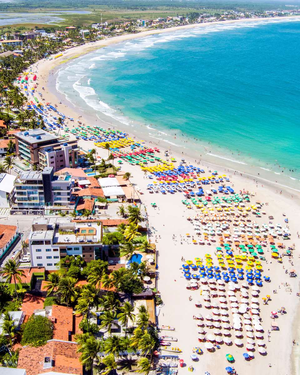 Praia de Porto de Galinhas vista do alto, com centenas de guarda-sóis coloridos na areia, o mar azul-turquesa, a vila de prédios baixos e o coqueiral ao fundo.