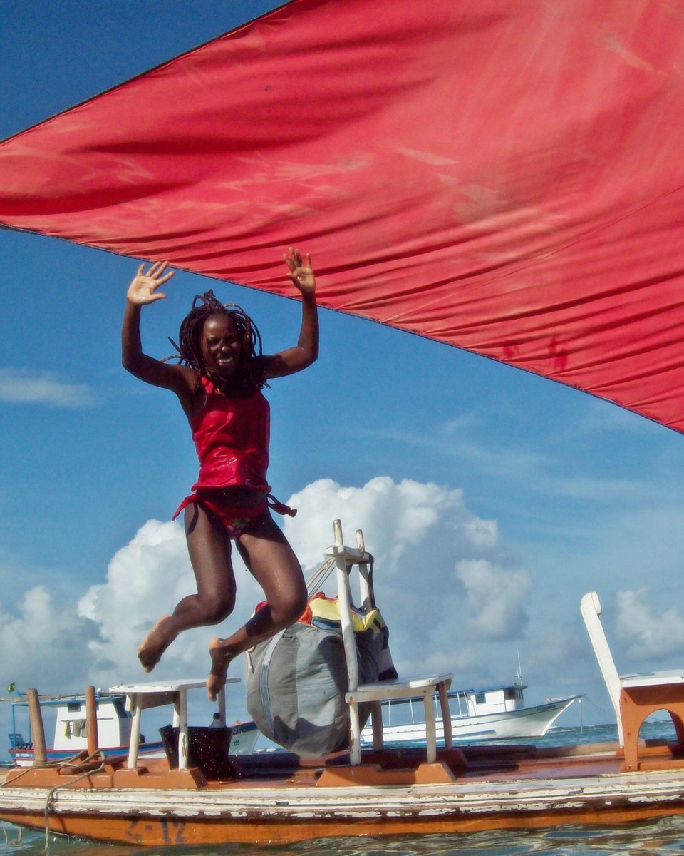 Mulher de roupa vermelha pulando de braços abertos sobre uma jangada de madeira, ao lado da vela vermelha, com o mar e as nuvens brancas ao fundo, em Porto de Galinhas.