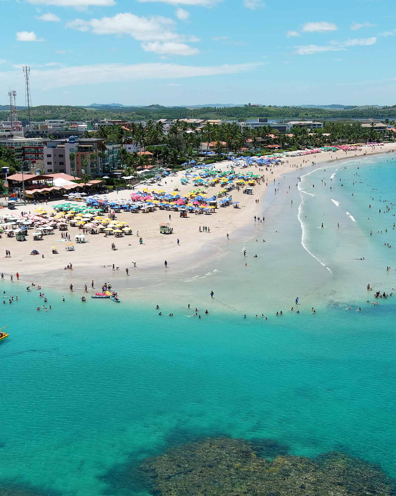Orla de Porto de Galinhas vista do alto num dia de sol, com a areia cheia de guarda-sóis coloridos, banhistas no mar verde e os recifes das piscinas naturais em primeiro plano.